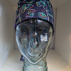May include: A black scrub hat with a neon green, blue and purple print of video game controllers and game consoles.