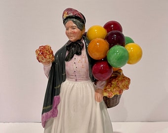 May include: A porcelain figurine of a woman wearing a black shawl and a white dress, holding a basket of flowers and a bunch of colorful balloons.