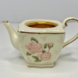 Puede incluir: Una tetera pequeña de porcelana blanca con cuerpo cuadrado y ribete dorado. La tetera tiene un diseño floral con rosas rosas y hojas verdes.