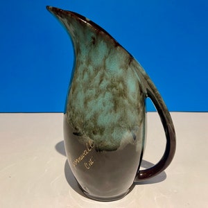 May include: A green and brown ceramic pitcher with a unique shape and a gold inscription that reads "Cornwall Ont."