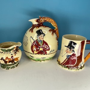 Może przedstawiać: Trzy ceramiczne kubki z motywem myśliwskim. Kubki są ozdobione obrazami ludzi w strojach myśliwskich i lisem. Kubki są w odcieniach brązu, kremowego i czerwonego.