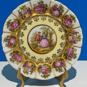 May include: A white porcelain plate with a gold rim and intricate gold designs. The center of the plate features a romantic scene of a couple in a garden setting. The plate is surrounded by 12 smaller scenes of couples in various settings, all with gold accents.