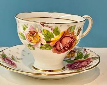 May include: A white teacup and saucer with a floral pattern featuring pink, yellow, and green roses. The cup and saucer have a gold trim.
