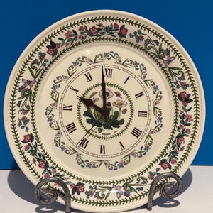 May include: A round ceramic wall clock featuring a cream-colored background with a floral and butterfly border pattern. The clock face displays black Roman numerals and dark brown hands, with a central botanical illustration and the text Portmeirion.