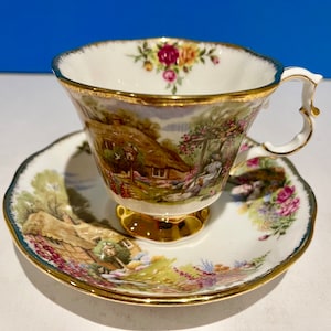Puede incluir: Un juego de taza y platillo de té blanco con borde dorado y estampado floral. La taza de té presenta una escena de cabaña con techo de paja y un jardín.