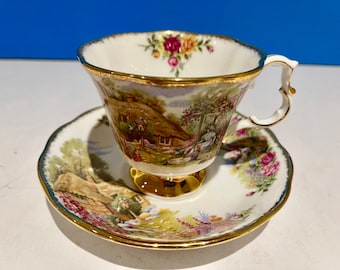 Puede incluir: Un juego de taza y platillo de té blanco con borde dorado y estampado floral. La taza de té presenta una escena de cabaña con techo de paja y un jardín.