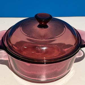 Puede incluir: Una fuente de horno de vidrio de color rubí vintage con tapa a juego. La fuente tiene dos asas laterales y una tapa con un pomo central. La tapa presenta un diseño radial. La fuente está hecha de vidrio transparente.