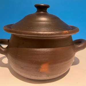 Peut inclure: Un pot en terre cuite brun avec un couvercle. Le pot a deux anses et est fait d'une argile rugueuse et texturée.