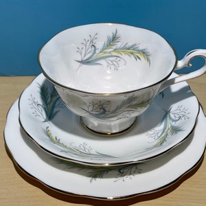 Puede incluir: Un juego de taza y platillo de té blanco con un delicado patrón floral en tonos grises y amarillos. El juego tiene un borde dorado.