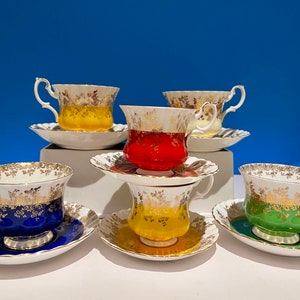 May include: A collection of six porcelain teacups and saucers in various colors including blue, red, yellow, green, and white. Each cup features a scalloped rim, a curved handle, and a decorative gold floral pattern around the upper edge.