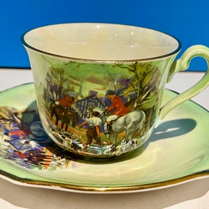 May include: A green and white teacup and saucer set with a gold trim. The set features a hunting scene with riders on horseback and hounds.