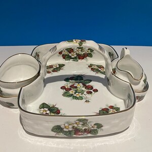 Puede incluir: Una bandeja de servir de porcelana blanca con asa, decorada con un patrón de fresas. La bandeja tiene dos cuencos pequeños y una jarra de crema.