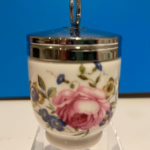 May include: A white porcelain egg cup with a floral design featuring pink, blue, and yellow flowers. The egg cup has a silver lid with a loop handle.