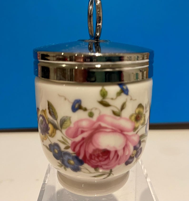 May include: A white porcelain egg cup with a floral design featuring pink, blue, and yellow flowers. The egg cup has a silver lid with a loop handle.