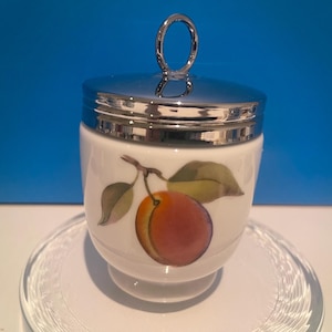May include: A white porcelain egg cup with a silver lid and a painted image of a peach with green leaves. The egg cup is sitting on a clear glass saucer.