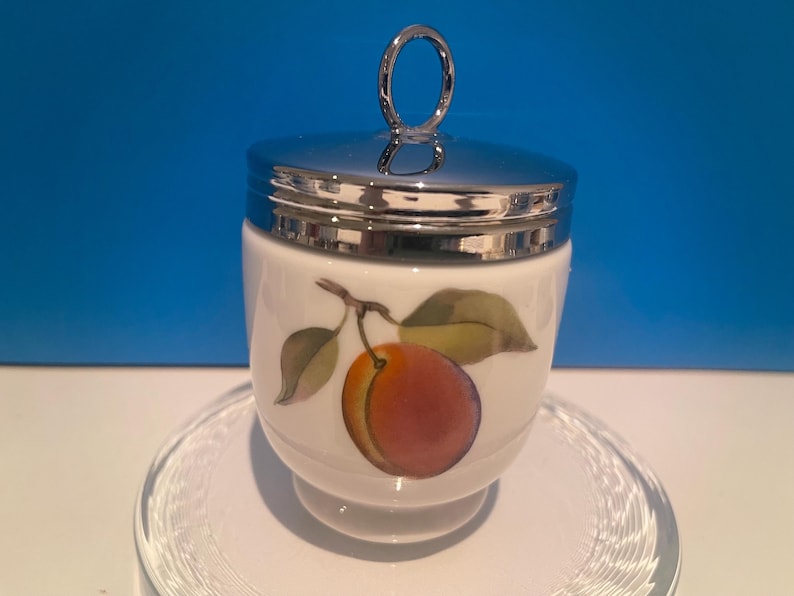 May include: A white porcelain egg cup with a silver lid and a painted image of a peach with green leaves. The egg cup is sitting on a clear glass saucer.
