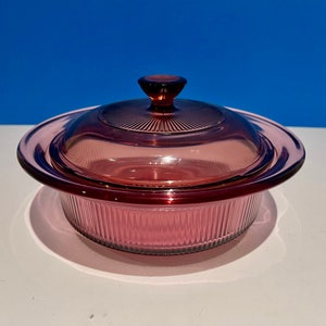 Puede incluir: Una cacerola de vidrio rosa vintage con tapa. La cacerola tiene un diseño acanalado y la tapa tiene un pomo en la parte superior.