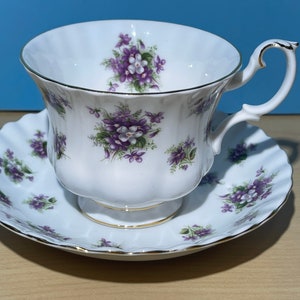 Puede incluir: Un juego de taza y platillo blanco con un patrón floral de violetas moradas. La taza tiene un borde dorado y el platillo tiene un borde festoneado.