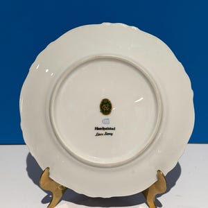 May include: White porcelain plate with a scalloped edge. The back of the plate has a green and gold logo with the text "Handpainted Love Story".