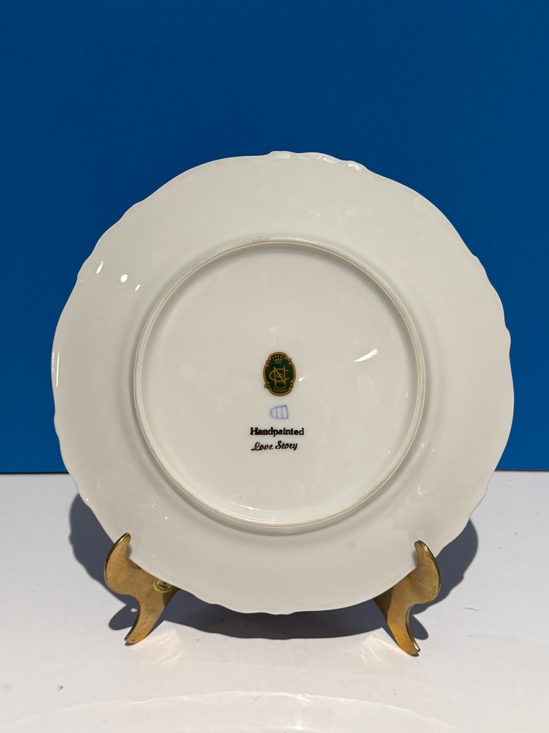 May include: White porcelain plate with a scalloped edge. The back of the plate has a green and gold logo with the text "Handpainted Love Story".