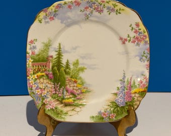 May include: A square, white ceramic plate with a gold rim and a floral design. The plate features a painted scene of a garden with a stone path, flowers, and trees. The plate is on a gold stand.