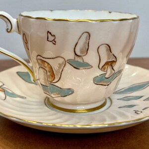 Puede incluir: Un juego de taza y platillo de color crema con un diseño de champiñones. La taza y el platillo tienen un borde dorado y están decorados con champiñones marrones y verdes.