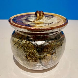 Pode incluir: Um pequeno pote redondo de cerâmica com tampa. O pote é decorado com um esmalte marrom, branco e verde. A tampa tem uma pega no topo.