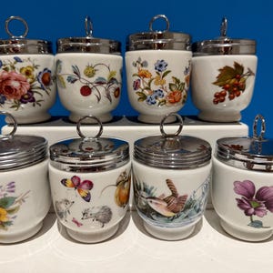 Op de afbeelding: Een set van acht witte porseleinen eierkokers met zilverkleurige metalen deksels met ringgrepen. Elke eierkoker toont verschillende botanische, florale, vlinder-, egel- of vogelillustraties in diverse kleuren, gerangschikt in twee rijen van vier.