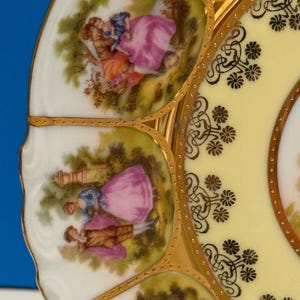 May include: A white porcelain plate with a yellow center and gold trim. The plate features a repeating pattern of stylized flowers and a scene of two people in a garden.