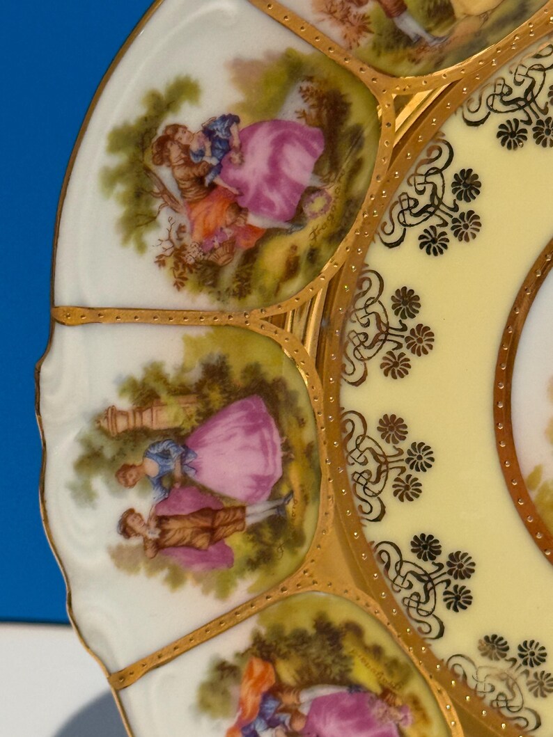 May include: A white porcelain plate with a yellow center and gold trim. The plate features a repeating pattern of stylized flowers and a scene of two people in a garden.
