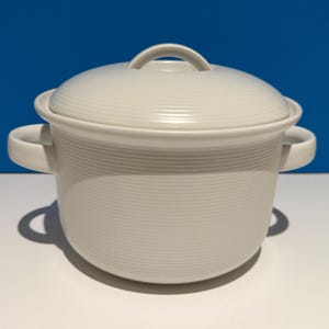 May include: A white ceramic pot with a matching lid, featuring a subtle horizontal ribbed texture throughout the exterior. The pot includes two curved side handles and a rounded lid handle, presenting a clean, uniform, and monochromatic design.