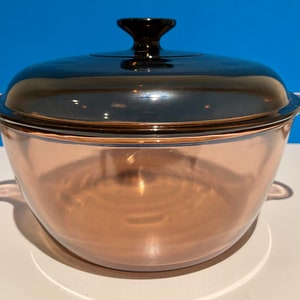 Puede incluir: Una fuente para horno vintage de vidrio transparente con tapa y asa de color marrón oscuro. El plato tiene forma redondeada y está diseñado para cocinar y servir. La tapa tiene un pomo negro para facilitar su manejo.