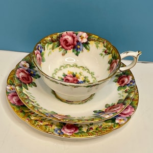 Peut inclure: Un ensemble de tasse et soucoupe en porcelaine blanche avec un motif floral dans des tons de rose, bleu et vert. L'ensemble est orné d'une bordure dorée et d'un bord festonné.