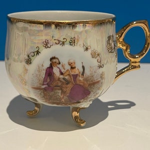 Puede incluir: Una taza de té de porcelana blanca con un borde dorado y tres patas doradas. La taza presenta una escena pintada de una pareja con ropa del siglo XVIII.