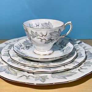 Könnte beinhalten: Ein Set aus vier weißen Porzellantellern mit einem silbernen und grauen Blattmotiv. Die Teller sind ineinander verschachtelt und haben einen goldenen Rand. Eine passende Teetasse und Untertasse befinden sich in der Mitte der Teller.