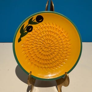 Peut inclure: Une assiette à râper en céramique jaune avec un bord vert et un motif de branche d'olivier. L'assiette a une surface texturée pour râper et est présentée sur un support doré. La branche d'olivier présente des olives noires et des feuilles vertes.