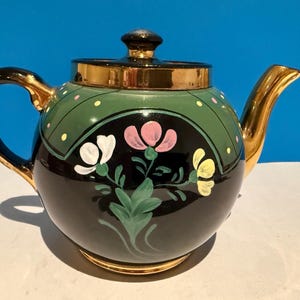 Peut inclure: Une théière ronde en céramique avec une base noire brillante ornée d'un motif floral peint à la main en blanc, rose et jaune. La partie supérieure est vert sauge avec de petits points jaunes et roses, complétée par une anse, un bec et un couvercle dorés.