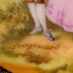 May include: A close-up of a porcelain figurine with a signature that reads "Fragonard". The figurine depicts a couple in a garden setting. The man is wearing a brown jacket and white stockings, and the woman is wearing a pink dress.
