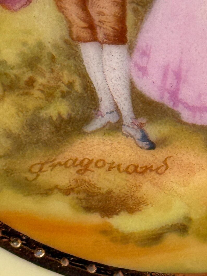 May include: A close-up of a porcelain figurine with a signature that reads "Fragonard". The figurine depicts a couple in a garden setting. The man is wearing a brown jacket and white stockings, and the woman is wearing a pink dress.
