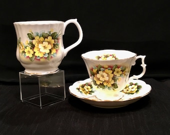 May include: A white teacup and saucer set with a floral design. The teacup has a handle and is decorated with yellow and green flowers. The saucer has a scalloped edge and matches the teacup's floral pattern. The teacup is on a clear acrylic stand.
