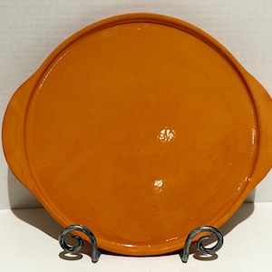 May include: A round, orange ceramic plate with two handles. The plate is displayed on a decorative metal stand. The plate has a glossy finish and is a solid, warm orange color. The plate is likely for serving food.
