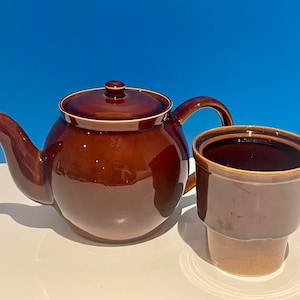 以下が含まれることがあります： 蓋付きの茶色の陶器製のティーポットと蓋付きの茶色の陶器製のマグカップ。ティーポットとマグカップは白い表面の上に置かれています
