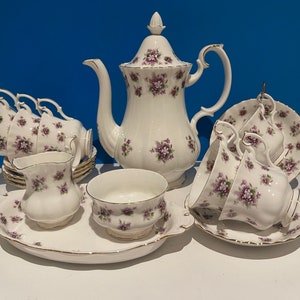 Puede incluir: Juego de té de porcelana blanca con un diseño floral morado y ribetes dorados. El juego incluye una tetera, tazas, platillos, una jarra de crema, una azucarera y una bandeja de servicio. El juego de té está dispuesto sobre una superficie blanca.