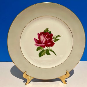 May include: A white porcelain dinner plate with a green and gold rim. The centre of the plate features a single red rose with green leaves.