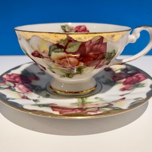 Puede incluir: Un juego de taza y platillo de té blanco con un diseño floral con rosas rosas, amarillas y verdes. El juego tiene un borde dorado.