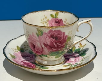 Puede incluir: Un juego de taza y platillo de té blanco con un patrón floral de rosas rosadas y hojas verdes. La taza y el platillo tienen un borde dorado.