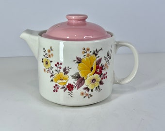 May include: A white ceramic teapot with a pink lid and floral pattern. The teapot has a handle and a spout. The floral pattern features yellow, red, and brown flowers.