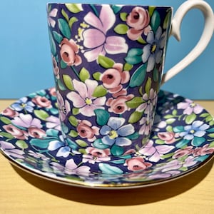 May include: A floral teacup and saucer set with a purple background and pink, blue, and green flowers. The cup has a white handle.