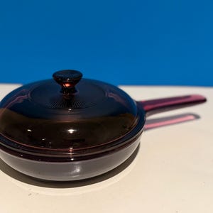 Könnte beinhalten: Eine dunkelviolette Glaspfanne mit passendem Deckel und Griff. Der Deckel hat ein kreisförmiges Design. Die Pfanne steht auf einer weißen Oberfläche mit blauem Hintergrund.