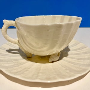 Puede incluir: Un juego de taza y platillo de té de porcelana blanca con diseño de concha. La taza tiene un asa y tres patas, mientras que el platillo tiene un borde festoneado.
